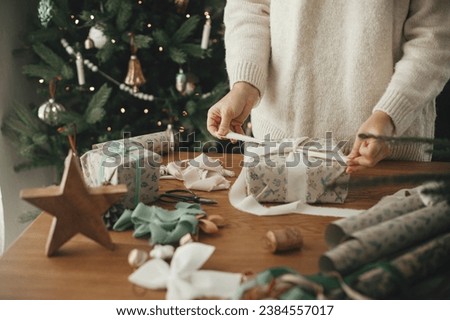 Woman in sweater wrapping christmas gift with stylish ribbon on wooden table with festive decorations in decorated scandinavian room. Merry Christmas! Hands packing present close up