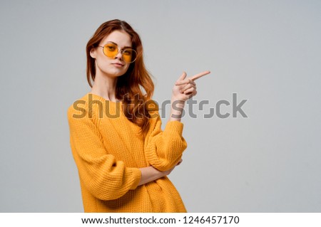 a woman in a sweater and yellow glasses shows a finger to the side                    