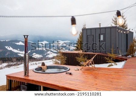 Woman swims in hot bath while resting at small modern house in the mountains on winter. Concept of recreation at tiny cabins in the mountains. Idea of escape to nature