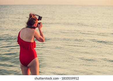 Woman In A Swimsuit With A Camera On The Sea, The Ocean. The Photographer Looks Into The Viewfinder Of The Camera. The Concept Of Summer Vacation, Freedom, Favorite Job, Profession. Copy Space