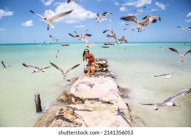 Woman in swimsuit stands on rocks next to the exotic sea laughing as flock of seagulls fly closely next to her.
