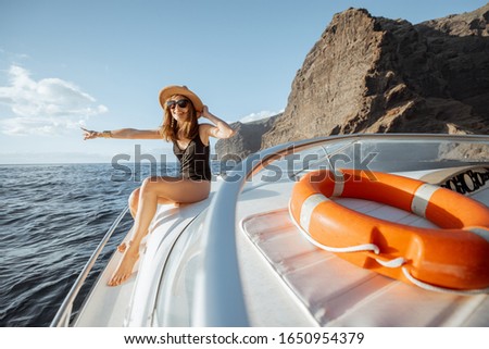 Woman in swimsuit and sun hat enjoying ocean voyage, sailing on a yacht near the breathtaking rocky coast on a sunset. Concept of a luxury summer recreational pursuit and travel