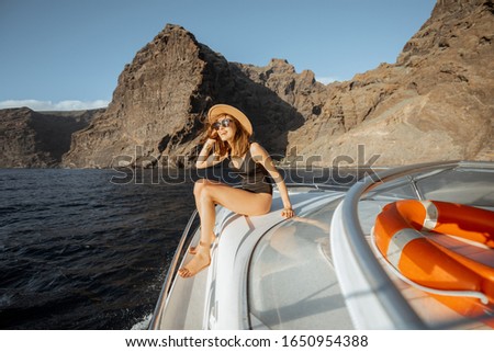 Woman in swimsuit and sun hat enjoying ocean voyage, sailing on a yacht near the breathtaking rocky coast on a sunset. Concept of a luxury summer recreational pursuit and travel