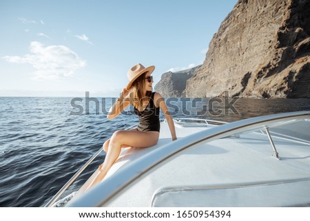 Woman in swimsuit and sun hat enjoying ocean voyage, sailing on a yacht near the breathtaking rocky coast on a sunset. Concept of a luxury summer recreational pursuit and travel
