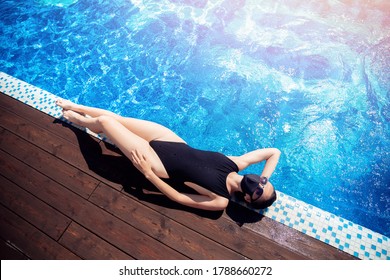 Woman In Swimsuit, Sunglasses And Protective Black Mask Sunbathes Against Backdrop Of Hotel Pool On Sun Lounger. Vacation Concept Covid Lifestyle.