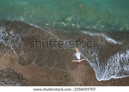 Woman with swimsuit and white dress resting on a sandy beach with braking waves on the shore. Overhead shot. Aerial drone photograph