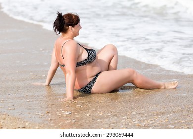 Woman in swimwear at the sea. Overweight young woman in swimsuit sits on wet sand in surf line