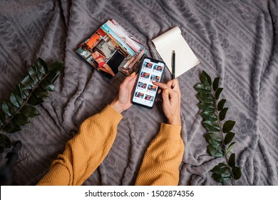 Woman Takes Cell Phone And Looks Photos On It. Mobile Phone, Notebook With Pen And Photos On The Soft Plaid. Flatlay With Mobile Phone, Photos And Notebook. Woman's Hands With Cell Phone.