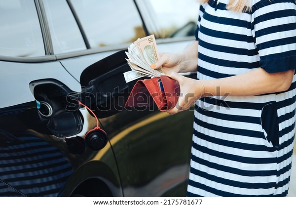 woman takes money dollars out of\
her wallet while standing next to a car with an open fuel\
tank