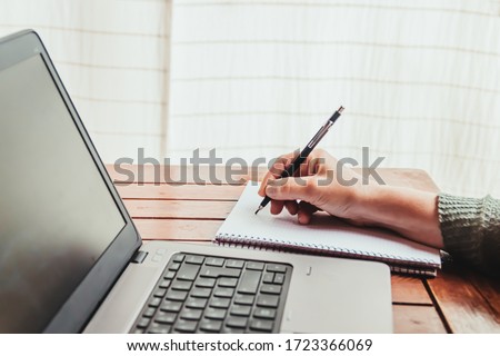 A woman takes notes on a paper next to a laptop. Concept of work from home during quarantine.