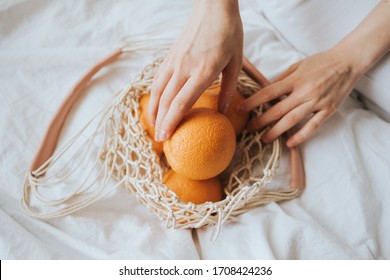 Woman Takes Oranges From A Net Eco Bag On A White Bed At Home