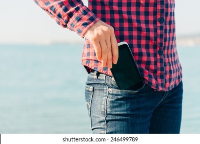 Woman takes out mobile phone of her pocket of jeans on beach near the sea to make self-portrait or to photograph the sea
