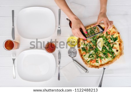 Woman takes a picture of a whole fresh pizza before eating on white wood table