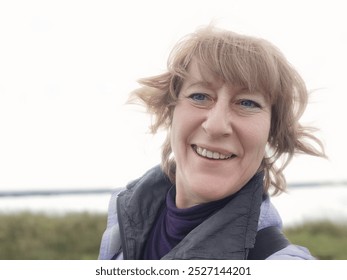 A woman takes a selfie with a cheerful smile, enjoying coastal breezes while documenting her travels as a blogger by the shoreline. - Powered by Shutterstock