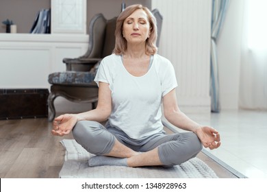 A Woman Takes Yoga At Home. Woman Over 50 Years Old. She Took The Pose Of The Asana On The Floor. Closing Her Eyes She Meditates. Close Up Shot.