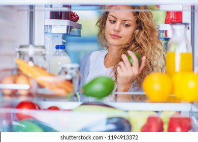 Woman Taking Avocado From Fridge To Prepare Breakfast While Standing In Front Of Opened Fridge. Picture Taken Form The Inside Of Fridge.