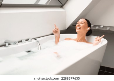 Woman Taking Bath With Foam Enjoying Relaxing Playlist Wearing Earbuds Earphones And Singing Listening To Music Online In Modern Bathroom At Home. Pampering And Relaxation - Powered by Shutterstock