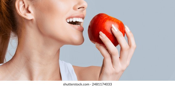 A woman is taking a bite out of a red apple, showing off her white teeth. The apple is held in her right hand, and the background is a soft grey. - Powered by Shutterstock