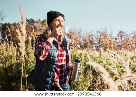 Woman taking break and relaxing with cup of coffee during summer trip. Woman standing on trail and looking away. Woman with backpack hiking through tall grass along path in mountains. Spending summer