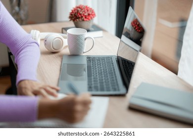 Woman taking notes in notebook while working or studying from home using laptop, headphones and coffee mug visible