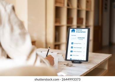 Woman taking notes while using a digital tablet to manage her home insurance policy, accessing overview, coverage details, payments, and claims