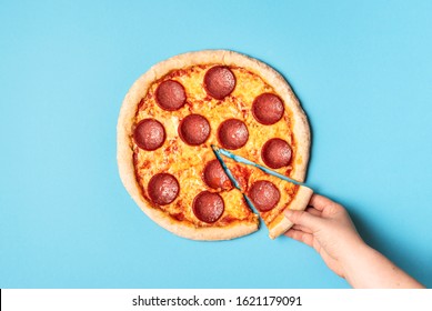 Woman Taking One Slice Of Pizza Salami On A Blue Background. Above View Of Pepperoni Pizza And A Hand Grabbing A Pizza Slice. Fast Food. Tasty Food.