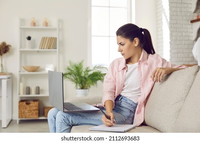 Woman Taking Online Course. Serious Focused Young Lady Sitting On Couch At Home, Watching Webinar On Laptop Computer, Using Modern Digital Learning Tools, Doing Research And Taking Study Notes