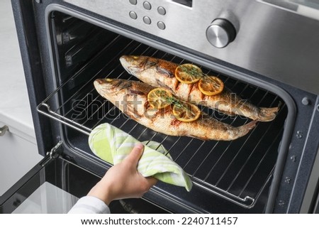 Woman taking out rack with sea bass fish, lemon and thyme from oven, closeup