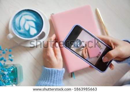 Woman taking photo of blue coffee latte, pink coloured planner 2021 and gold pen on her smartphone. People and technology. Taking picture for posting and sharing on social media. Blogging concept.