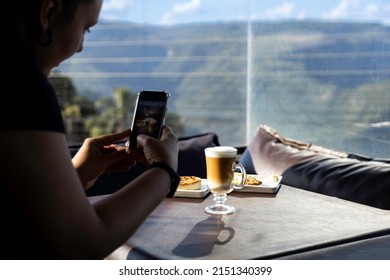 A woman taking photos of a latte macchiato coffee with a mountain view outdoors - Powered by Shutterstock