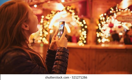 Woman Taking Pictures of European Christmas Market Scene on Smartphone. 4K. Girl Enjoying Winter Holiday Season, visiting Outdoors Christmas Market, Making photos on cell phone. Travel Europe