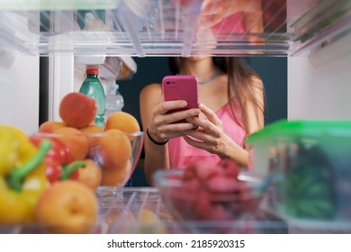 Woman Taking Pictures Of Food In The Fridge Using Her Smartphone, POV Shot From Inside Of The Fridge