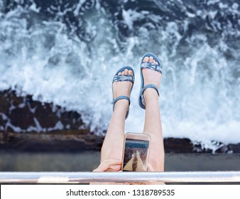 Woman taking pictures her feet with smart phone against the blue sea background. Womans slender legs in summer shoes top view