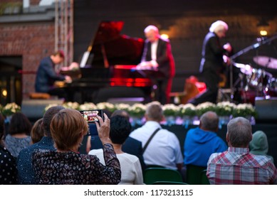 Woman Is Taking Pictures Or Recording Video Of Jazz Concert. Male Musicians On Stage Singing, Playing Piano, Saxaphone, Drums, Cello. Crowded Venue. Audience Enjoying Romantic And Relaxing Evening