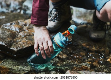 Woman Taking Pure Water To Bottle From Mountain Stream During Trekking In Mountains. Hiker Crouching On Rocks, Filling Bottle Up With Cold Mountain Water. Enjoying The Outdoors In The Summer Trip