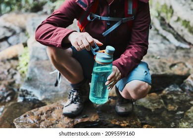 Woman taking pure water to bottle from mountain stream during trekking in mountains. Hiker crouching on rocks, filling bottle up with cold mountain water. Enjoying the outdoors in the summer trip