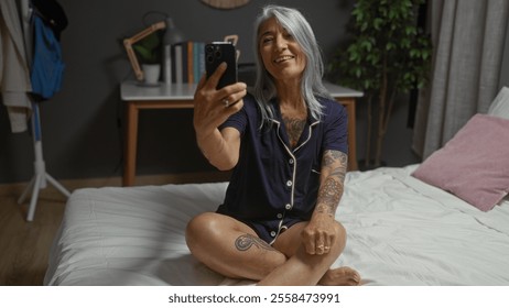 Woman taking selfie in a cozy bedroom while sitting on a bed, beautiful, grey-haired, mature, smiling, indoors, tattooed, casually dressed, and relaxed at home - Powered by Shutterstock