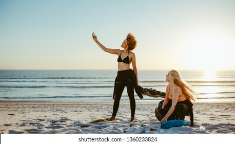 Woman Taking Selfie With Her Friend While Cleaning The Beach Area. Two Girls Tidying Up Rubbish On Beach And Taking A Selfie.