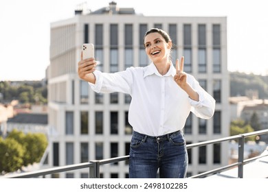 Woman taking selfie on city rooftop making peace sign, wearing white shirt and blue jeans, smiling and enjoying sunny day. Urban backdrop with modern buildings. - Powered by Shutterstock