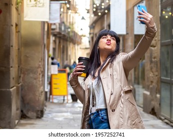 Woman Taking A Selfie On The Street Kissing The Cellphone Camera And Holding A Disposable Coffee Cup. Influencer Girl Being Silly On A Photo For Social Media Sharing. Brunette In A City Having Fun.