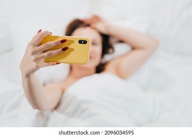 Woman taking selfie with smartphone while lying in comfortable bed. Lady holding mobile in light cozy bedroom. Addiction to technology and social media, portrait concept. High quality photo