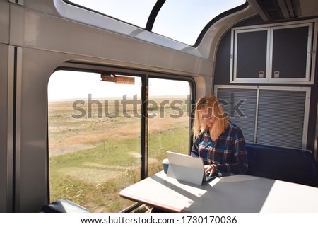 Woman taking train across USA, sitting at the table, using laptop  and drinking coffee. Inside lounge cabin with big windows. Rural scene outside.