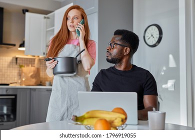 woman talk on phone while husband work on laptop at home, black guy look at talking woman, she holds saucepan in hands