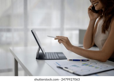 A woman is talking on her cell phone while using a laptop. She is smiling and she is in a good mood. The laptop is open and the woman is pointing at something on the screen. There are also a few pens