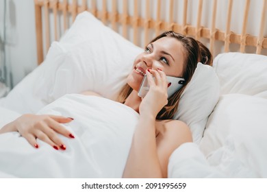 Woman talking on smartphone while lying in comfortable bed. Lady holding mobile in light cozy bedroom. Addiction to technology, morning rituals concept.