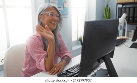 Woman talking on a videoconference call in an office setting, smiling confidently at the camera, wearing glasses and a pink shirt, with grey hair, surrounded by modern office equipment. - Powered by Shutterstock