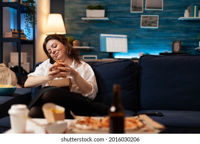 Woman talking at phone with friends while holding tasty delicious burger in hands relaxing on couch in living room late at night. Caucasian female enjoying tajeaway junk food home delivered - Powered by Shutterstock