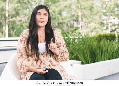 Woman Talking To Someone. Young Woman Smiling, Sitting On A Chair In The Mall, In A Green Area. Portrait Of A Colombian Girl Looking At The Camera. Woman Dressed In Jeans, Blouse And Tennis Shoes.