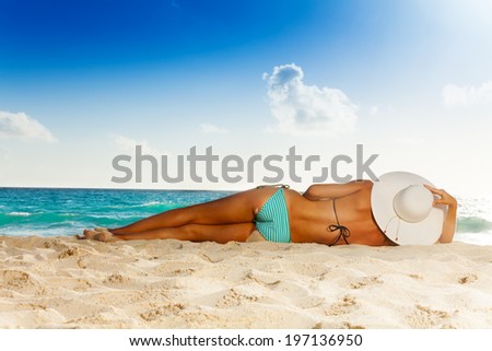  woman with tan laying on white sand beach wearing striped swimwear and big hat