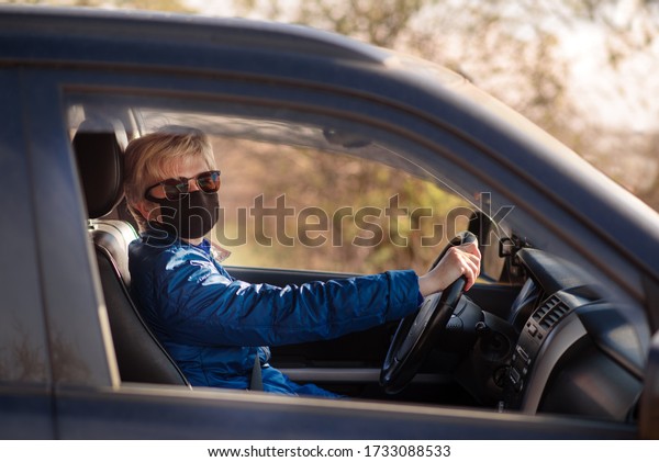 Woman taxi driver in face mask. Woman in black face mask\
and sunglasses is driving a car and lookiing in the driver\'s opened\
window. Left-hand drive. Coronavirus infection control concept.\
